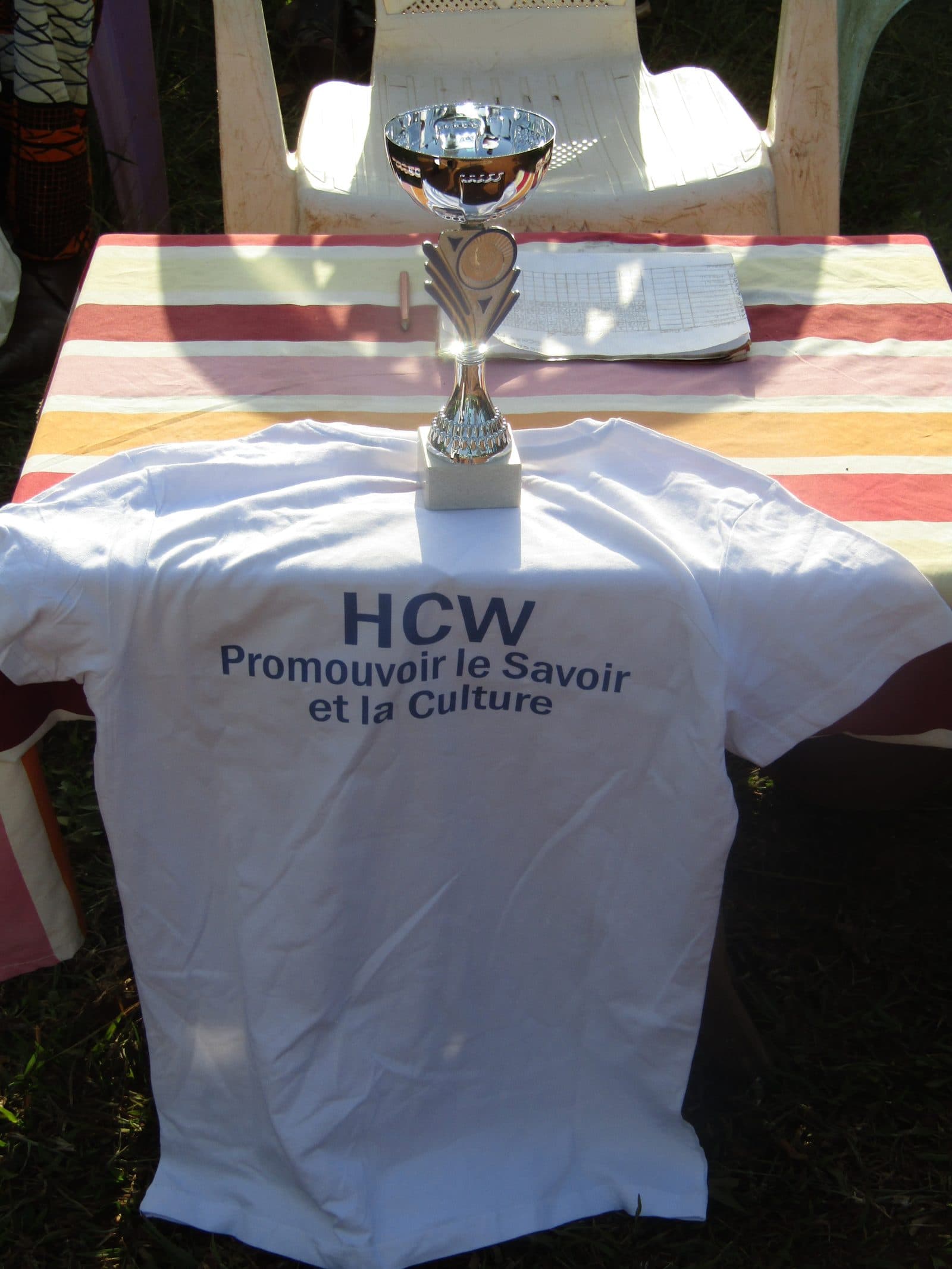 Un maillot blanc portant « HCW — Promouvoir le Savoir et la Culture », posé devant une coupe sur une table nappée.