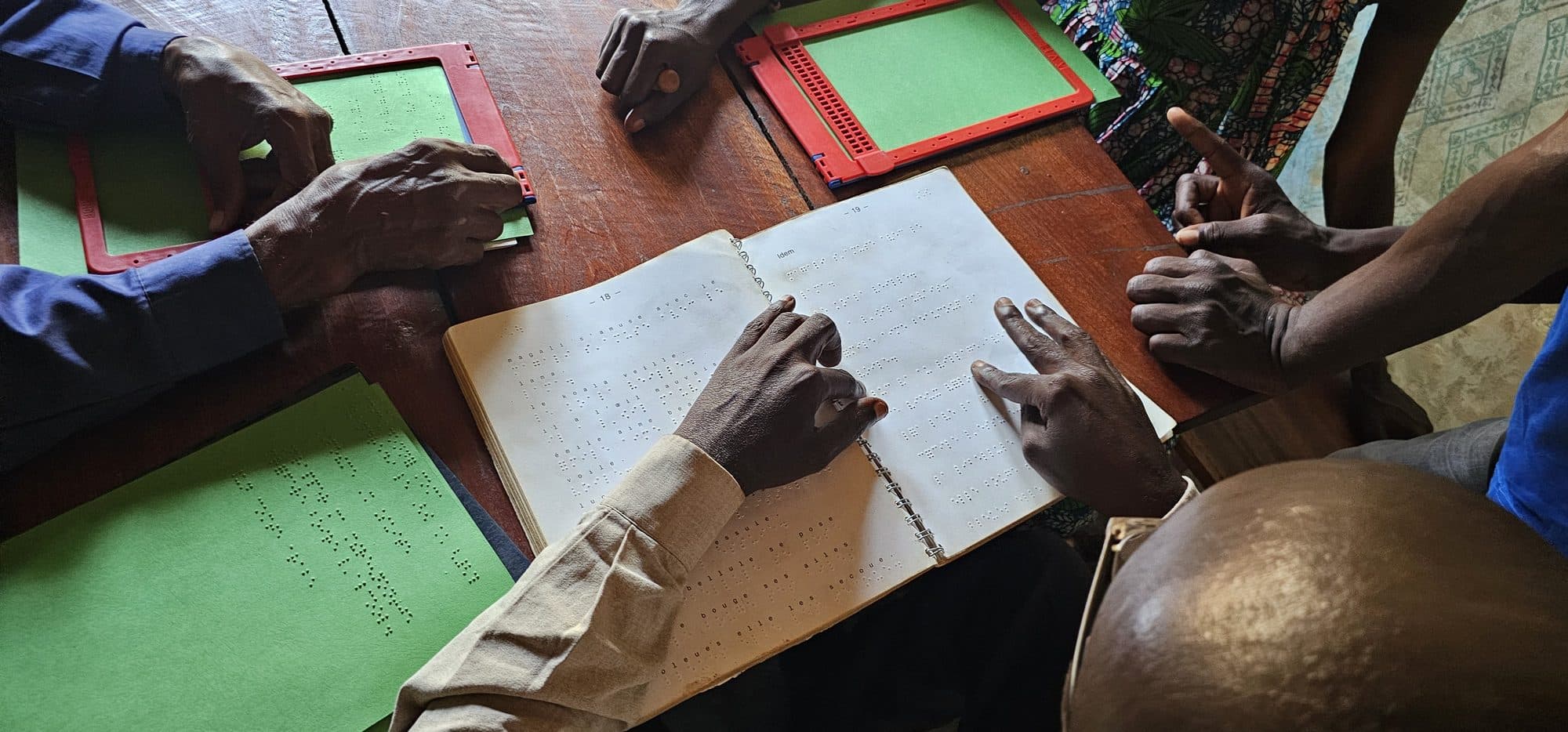 Des mains lisent un volume en braille posé sur une table ; à côté, des tablettes d'écriture et un poinçon.