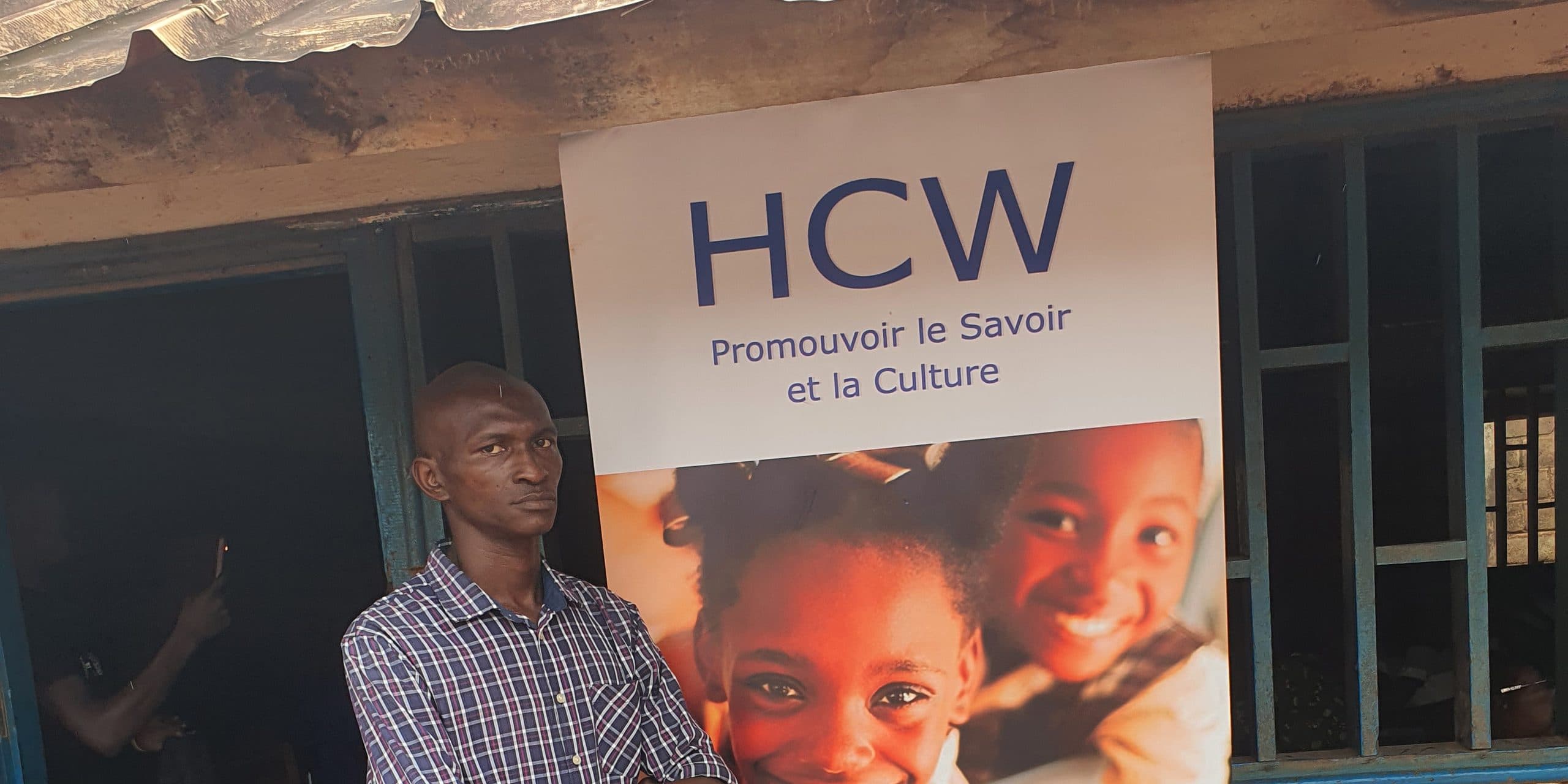 An HCW pull-up banner in front of a school building, reading “HCW — Promouvoir le Savoir et la Culture”. A man is standing beside it.