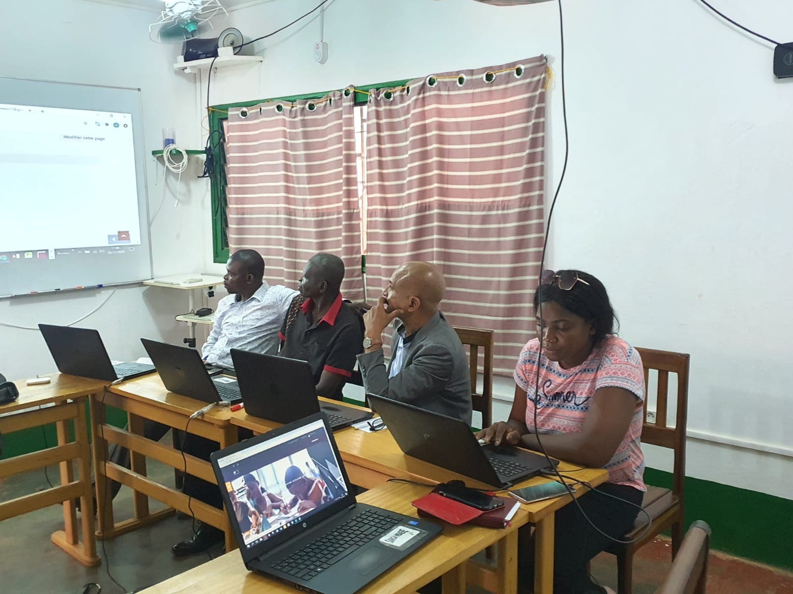 Quatre adultes assis en rang devant des ordinateurs portables dans une salle de formation ; trois regardent un écran de projection, une femme tape sur son clavier.