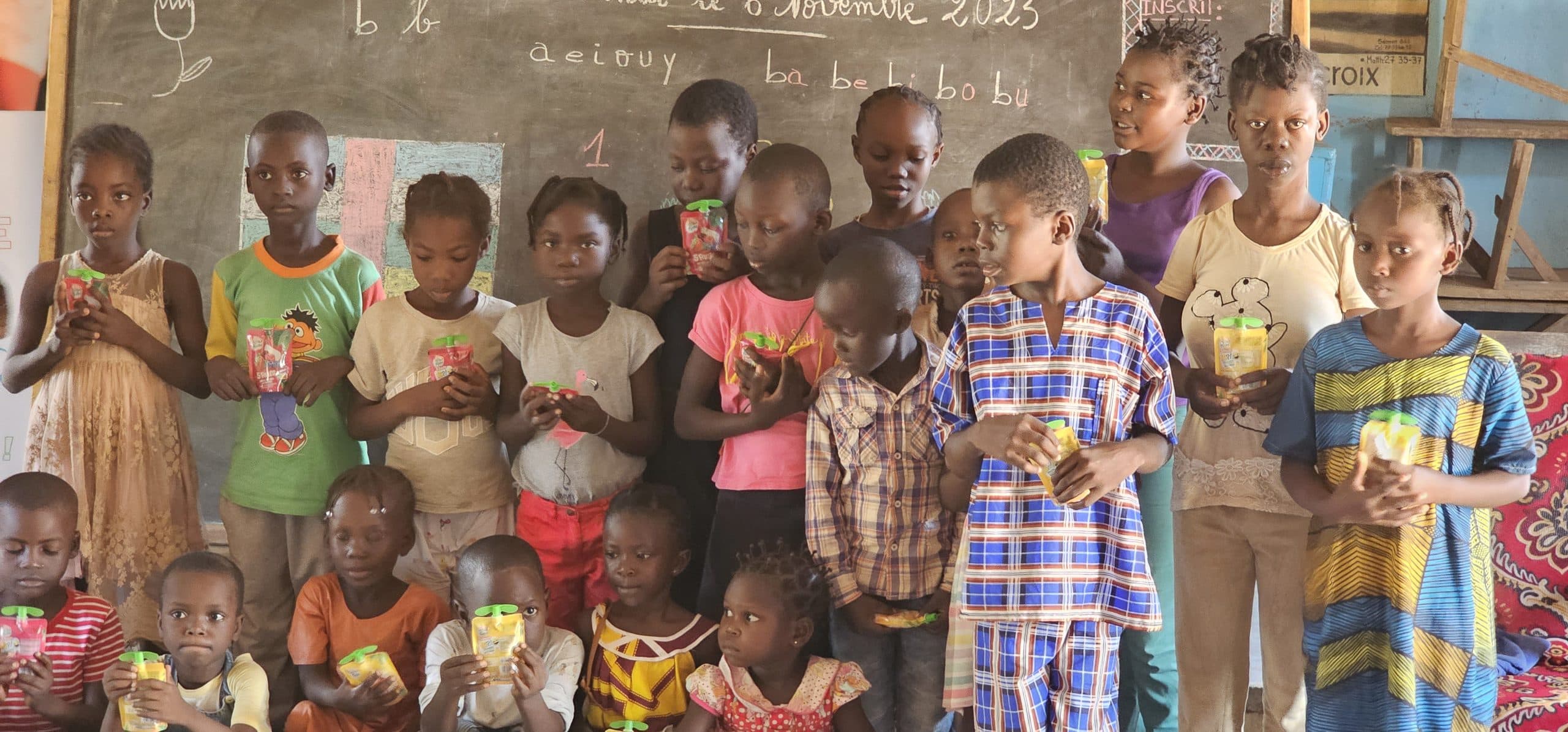 Un groupe d'enfants debout devant un tableau noir portant la date et un syllabaire ; chacun tient une petite boisson.